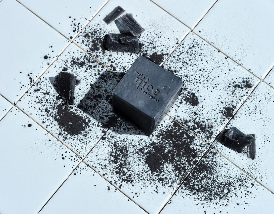 Black charcoal solid dish soap block in the center of light blue tile background, with scattered charcoal pieces and powders surrounding it.