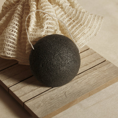 Charcoal Konjac Sponge on wooden plank and with natural showering pompom. Neutral minimal beige background.