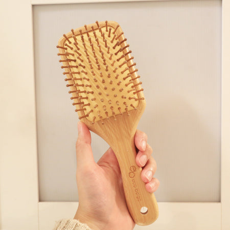 Rectangular cushioned bamboo hair brush with Eco Beige engraved on the handle. Hand held in minimal background.