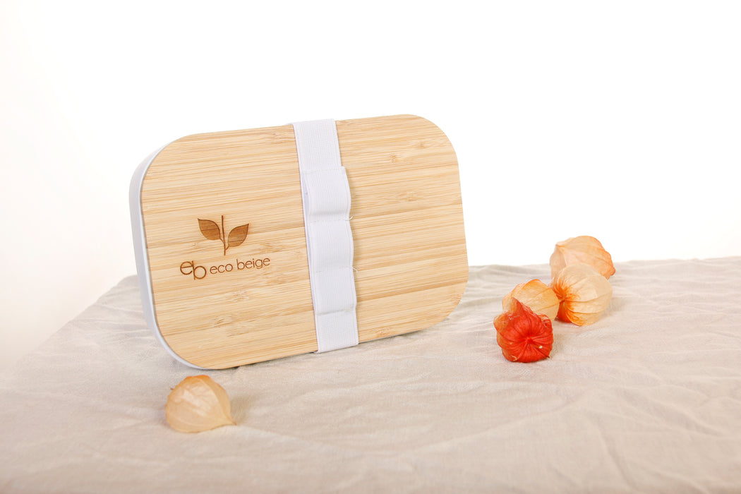 Eco Beige bamboo lunch box with engraved logo on the lid. Lunch box stands up with front view. White container made with wheat fiber and bamboo fiber. Comes with an elastic band for utensils. White background with linen table cloth and red gooseberry flowers.