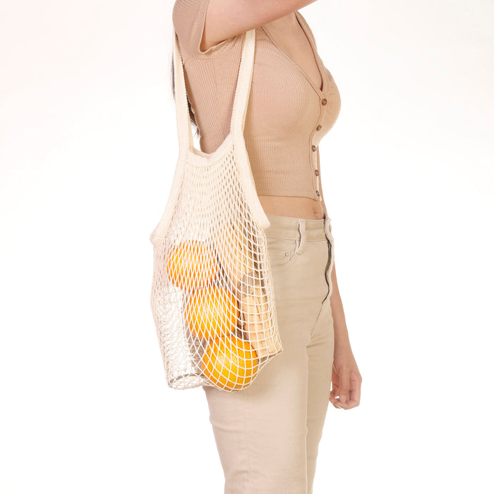 Eco Beige cotton netted shoulder bag with bottle, oranges, and pouch inside. Wore on shoulder by model in beige color theme outfit and white background.