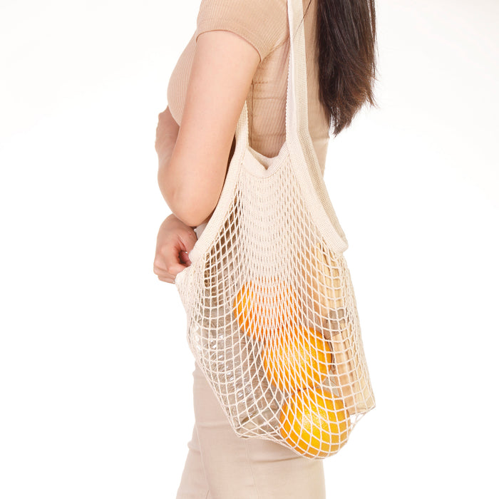 Eco Beige cotton netted shoulder bag with bottle, oranges, and pouch inside. Wore on shoulder by model in beige color theme outfit and white background.