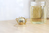 Mini Gold Teapot Tea Infuser sitting on wood surface with chain draping down beside it and a jar of tea on the side.