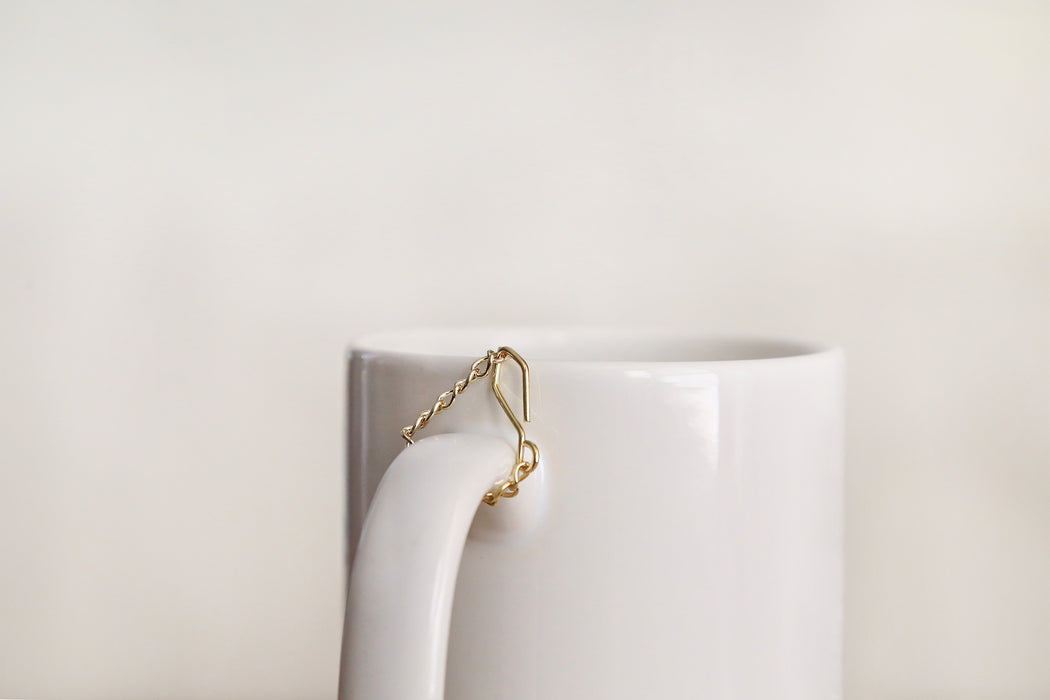Minimal clean background with tea infuser's gold chain hooked on a white mug handle.