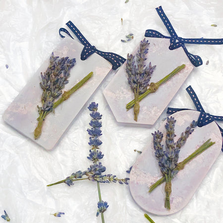 3 shapes of neutral toned purple plaster tablet fresheners with sprinkles of light pink and dried lavender flower decorations on top. Long hexagon, rectangle, and ellipse shape with ribbon tied at the top of tablet, used as a decorative diffuser. White tissue crumble background with lavender peices..