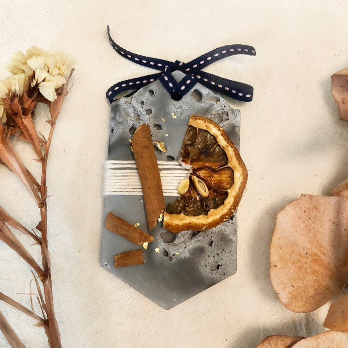 Musky Fall Plaster Freshener in grey concrete style and irregular texture. Decorated with dried citrus and cinnamon sticks. Long Hexagon shape with ribbon tied at the top of tablet, used as a decorative diffuser. Dried preserved brown plants at the background.