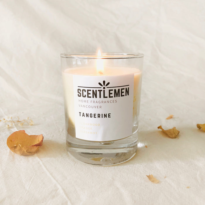 Scentlemen Studio hand poured soy wax tangerine scent candle in a glass container. Cream crumbled background with dried flower petals.
