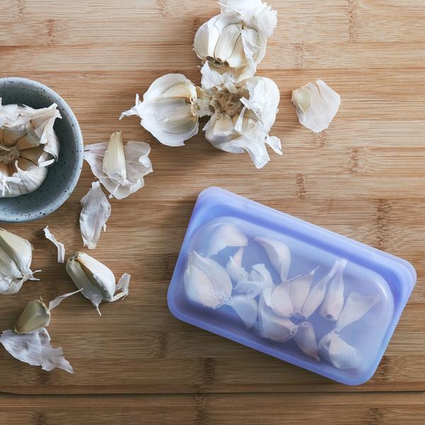 Stasher platinum silicone reusable amethyst color snack bag storing garlics. On wood counter with scattered garlic pieces.