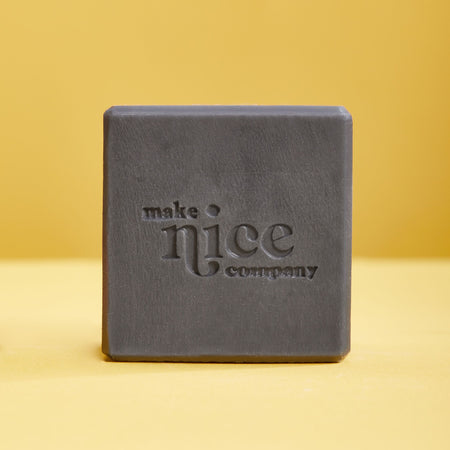 Black charcoal solid dish soap block with Make Nice Company logo pressed into the block. Contrasting yellow background color.